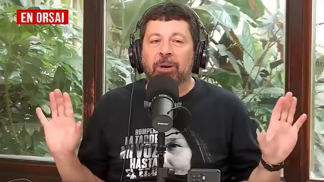 El economista Alfredo Zaiat, con auriculares y micrófono, gesticula mientras habla en su programa, con el logo de "EN ORSAI" en la esquina superior izquierda. Ilustra la crítica a la gestión económica de Luis Caputo.